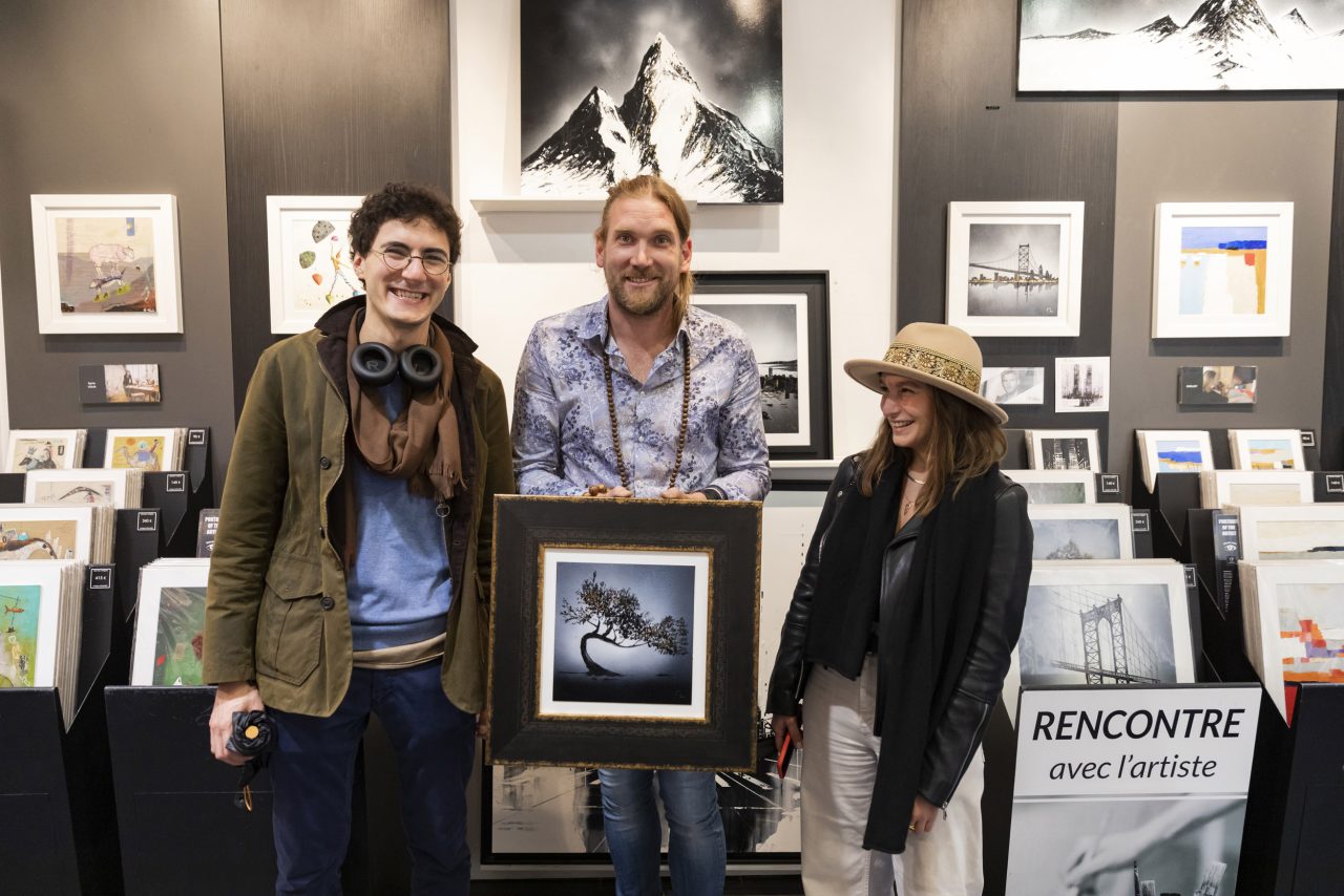 Portrait du peintre Julien Rey un tableau à la main au milieu d'un couple d'acheteur dans une galerie d'art 